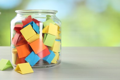 Photo of Colorful paper pieces in glass jar on wooden table against blurred background, closeup. Space for text
