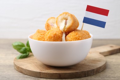 Photo of Delicious fried croquettes in bowl, flag of Netherlands and basil on wooden table, closeup