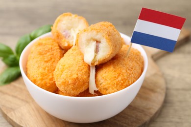 Photo of Delicious fried croquettes in bowl, flag of Netherlands and basil on wooden table, closeup