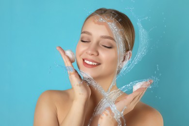 Image of Young woman washing face with cleansing foam and water splashes on light blue background