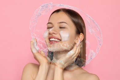 Image of Young woman washing face with cleansing foam and water splash on pink background