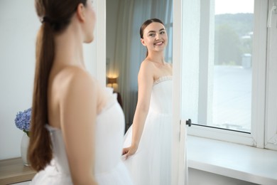 Photo of Dreams come true. Woman wearing beautiful wedding dress looking at herself in mirror indoors
