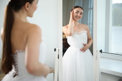 Photo of Dreams come true. Woman wearing beautiful wedding dress looking at herself in mirror indoors