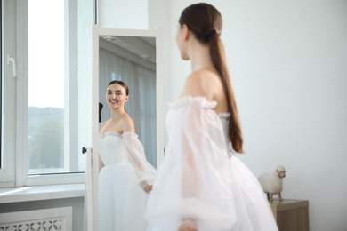 Photo of Dreams come true. Woman wearing beautiful wedding dress looking at herself in mirror indoors