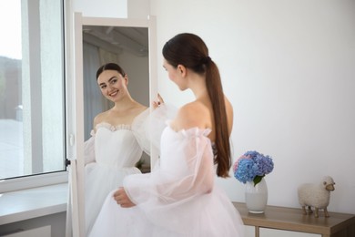 Photo of Dreams come true. Woman wearing beautiful wedding dress looking at herself in mirror indoors