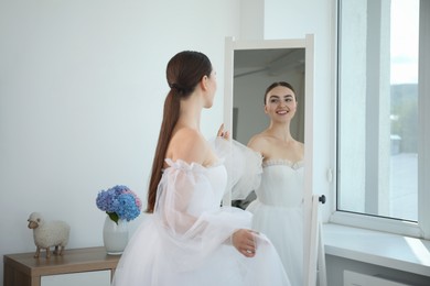 Photo of Dreams come true. Woman wearing beautiful wedding dress looking at herself in mirror indoors