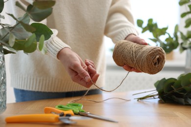 Photo of Florist with spool of thread making beautiful bouquet of eucalyptus branches at wooden table in flower shop, closeup