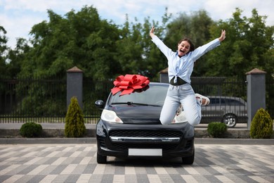 Photo of Dreams come true. Woman jumping near her new auto with big red bow outdoors