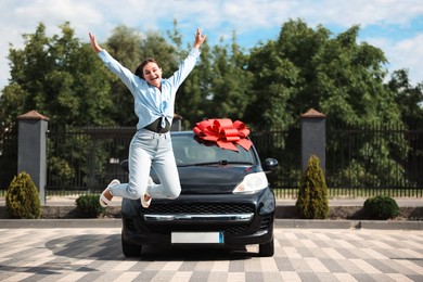 Photo of Dreams come true. Woman jumping near her new auto with big red bow outdoors