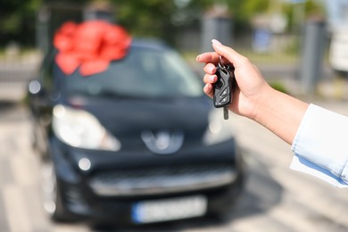 Photo of Dreams come true. Woman holding car key near her new auto with big red bow outdoors, closeup
