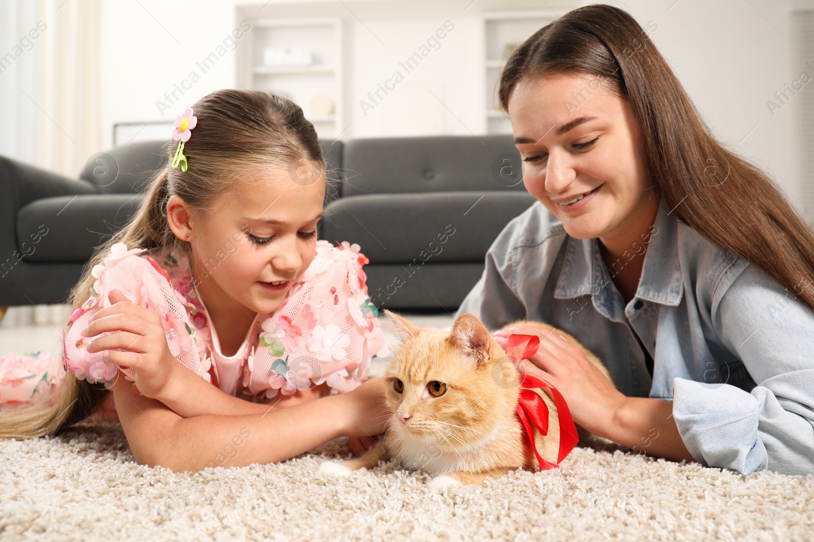 Photo of Dreams come true. Mother and her daughter stroking ginger cat with red bow on floor at home