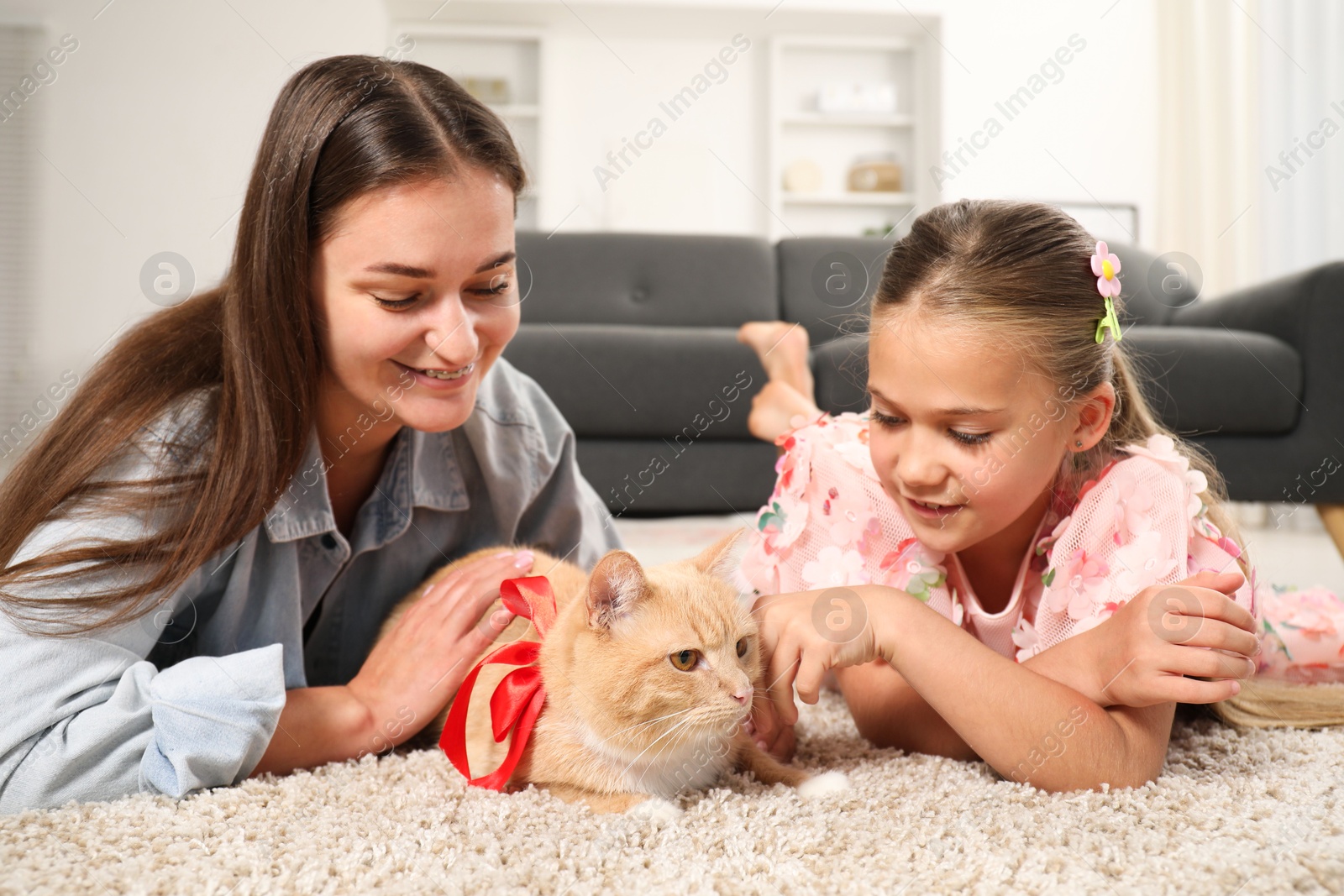 Photo of Dreams come true. Mother and her daughter stroking ginger cat with red bow on floor at home