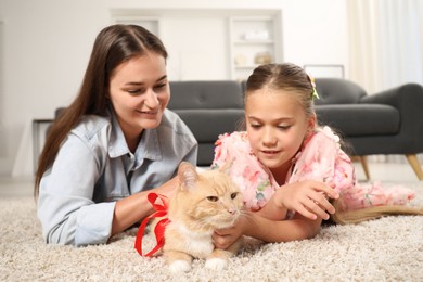 Photo of Dreams come true. Mother and her daughter stroking ginger cat with red bow on floor at home