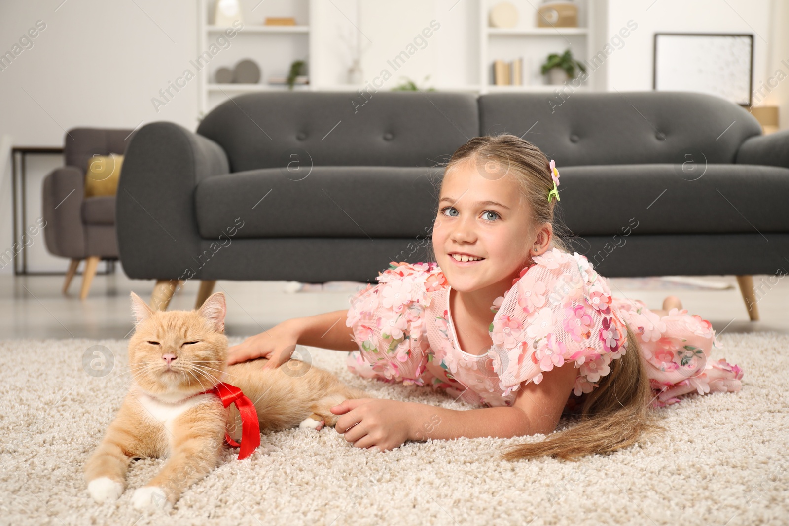 Photo of Dreams come true. Cute little girl stroking ginger cat with red bow on floor at home