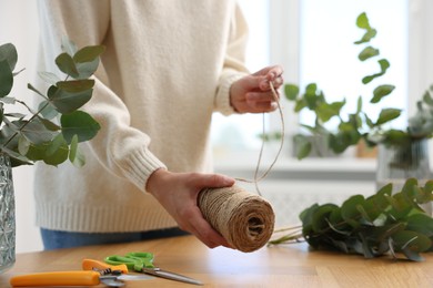Photo of Florist with spool of thread making beautiful bouquet of eucalyptus branches at wooden table in flower shop, closeup