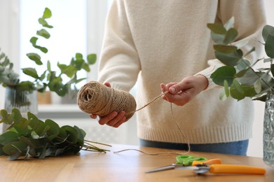Photo of Florist with spool of thread making beautiful bouquet of eucalyptus branches at wooden table in flower shop, closeup