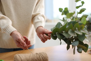 Photo of Florist with spool of thread making beautiful bouquet of eucalyptus branches at wooden table in flower shop, closeup