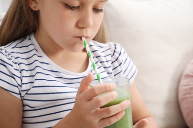 Photo of Little girl drinking juice through straw on sofa at home, closeup