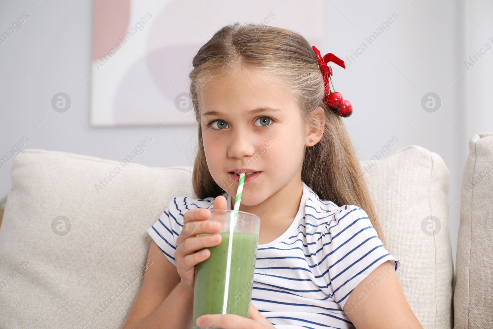 Photo of Little girl drinking juice through straw on sofa at home