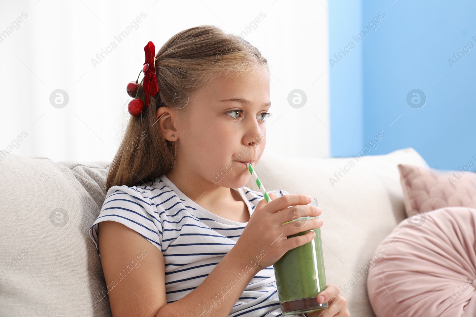 Photo of Little girl drinking juice through straw on sofa at home