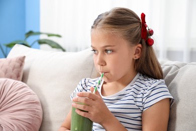 Photo of Little girl drinking juice through straw on sofa at home