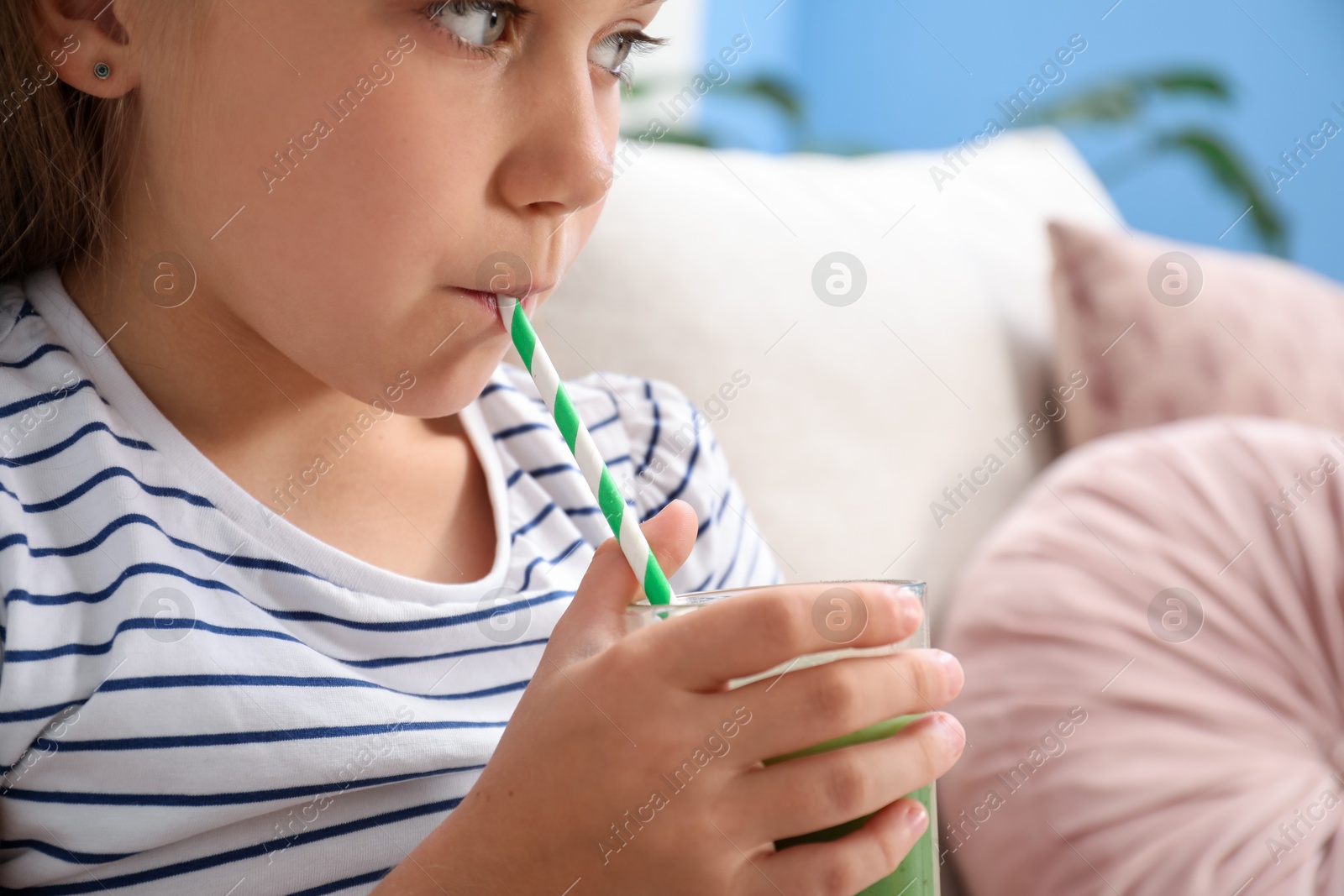Photo of Little girl drinking juice through straw on sofa at home, closeup