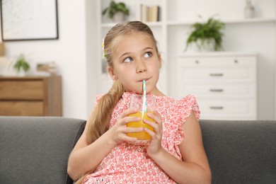 Photo of Little girl drinking juice through straw on sofa at home