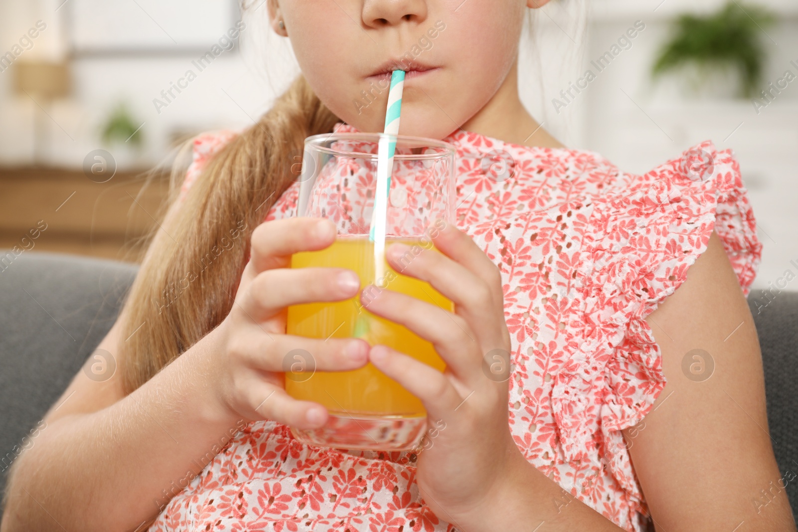 Photo of Little girl drinking juice through straw on sofa at home, closeup