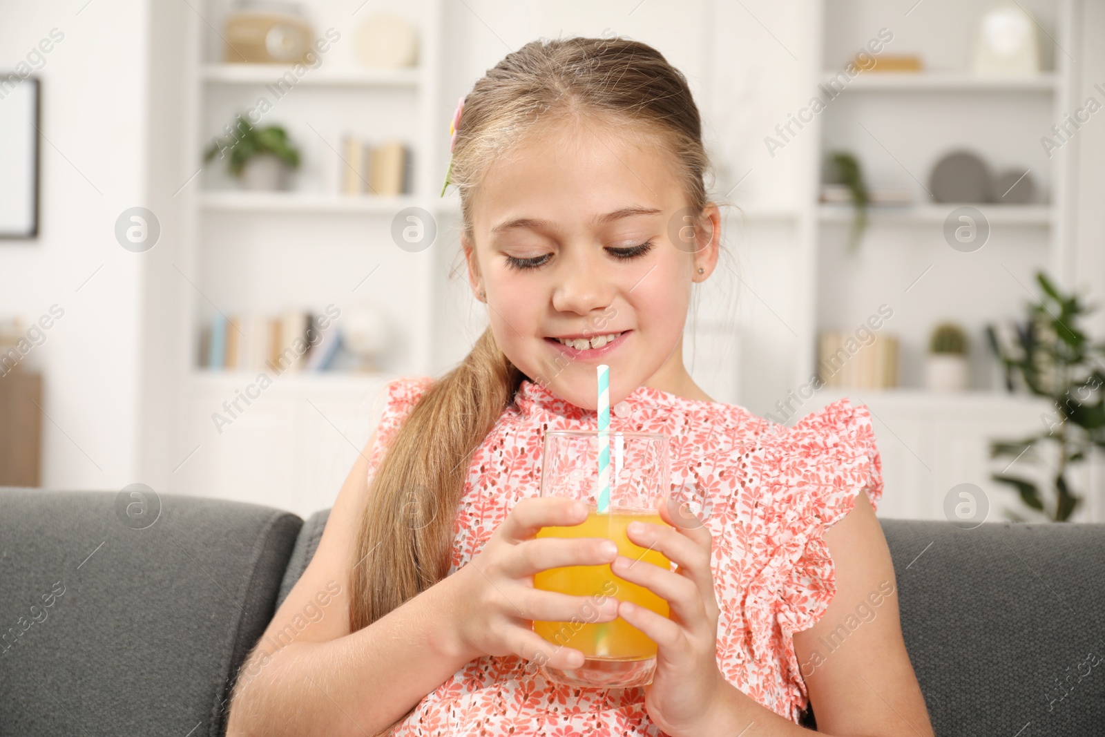 Photo of Little girl with juice and drinking straw on sofa at home