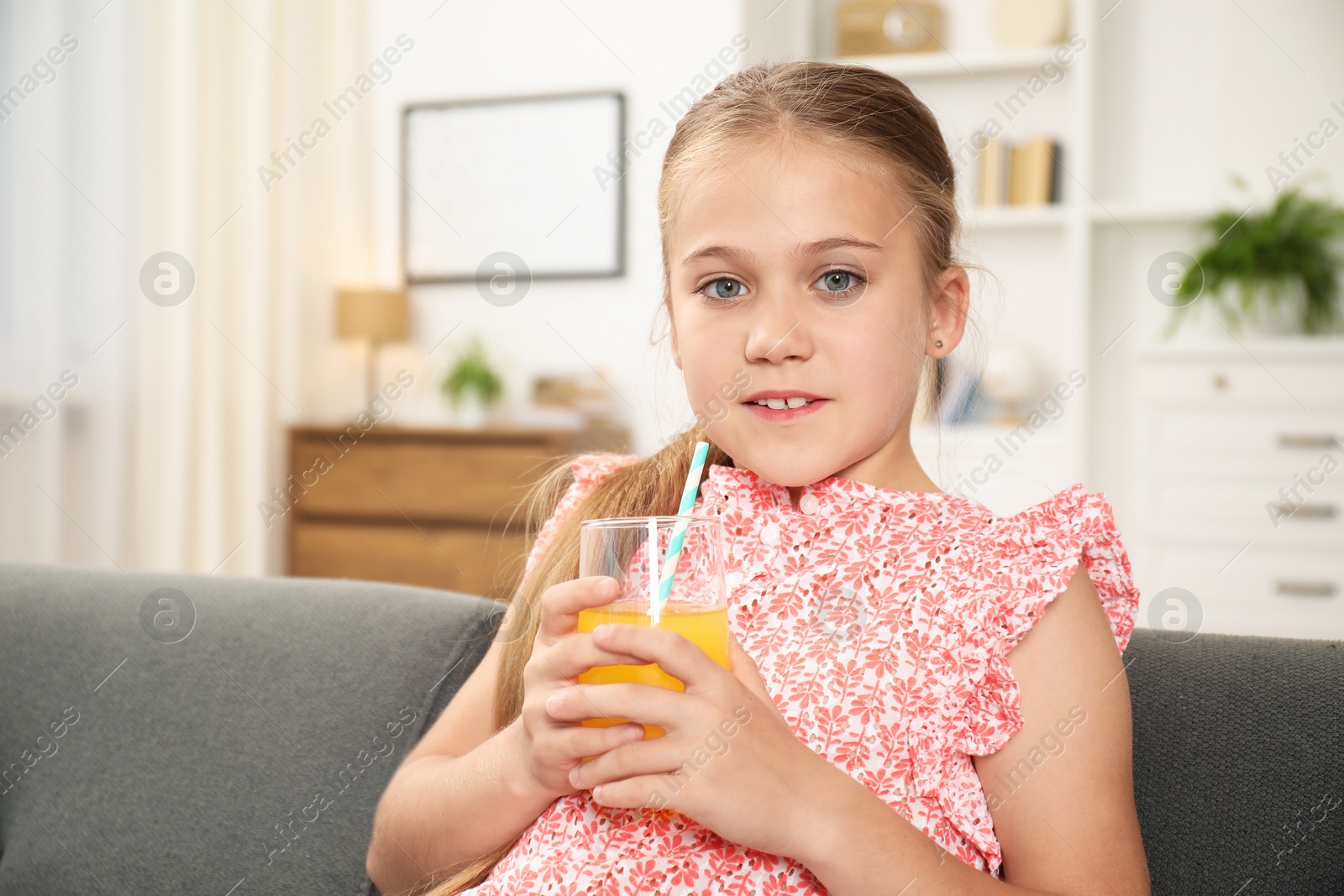 Photo of Little girl with juice and drinking straw on sofa at home