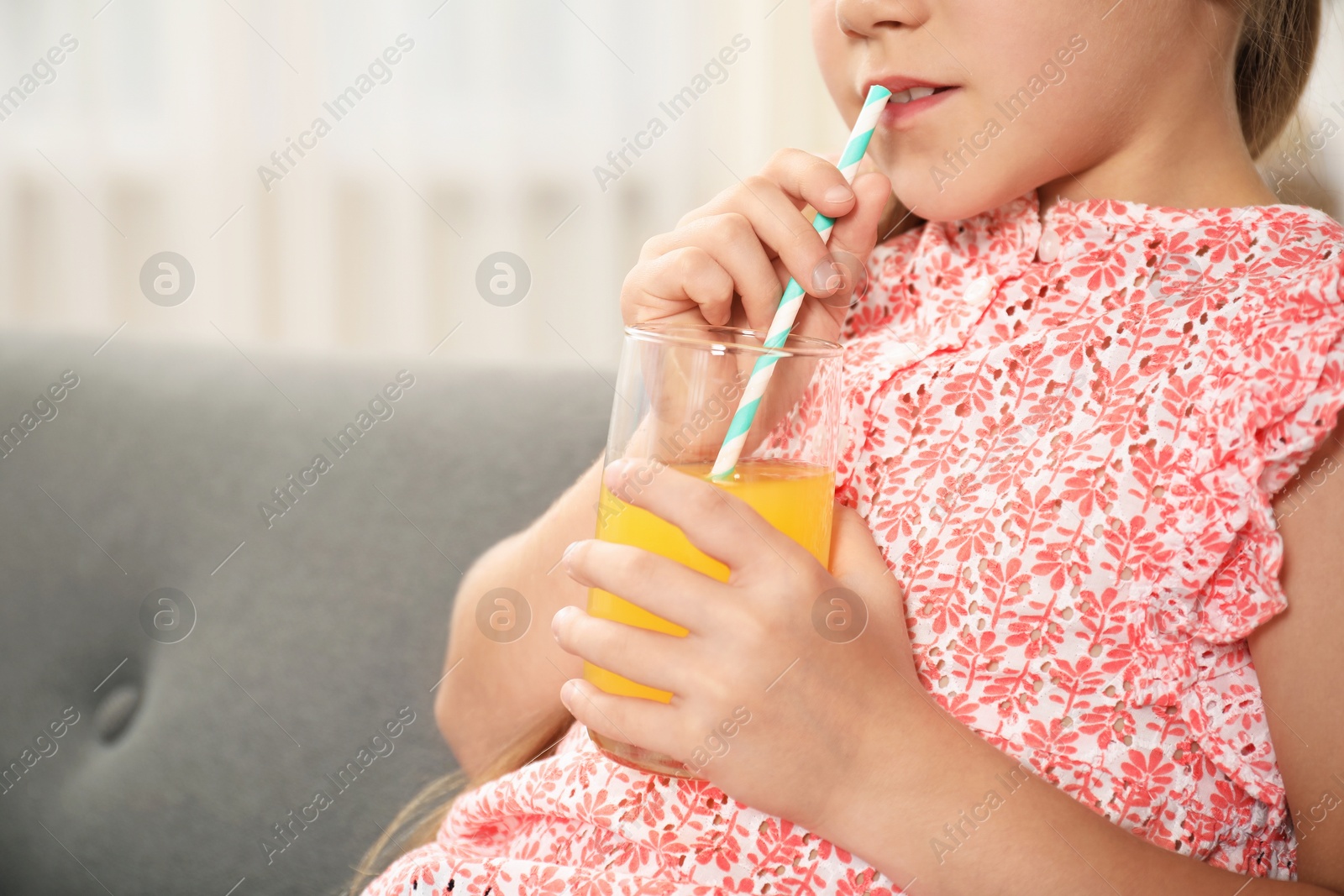 Photo of Little girl drinking juice through straw on sofa at home, closeup. Space for text