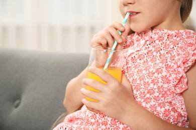 Photo of Little girl drinking juice through straw on sofa at home, closeup. Space for text