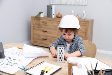 Photo of Choice of profession. Little kid in hard hat exploring house model at table indoors