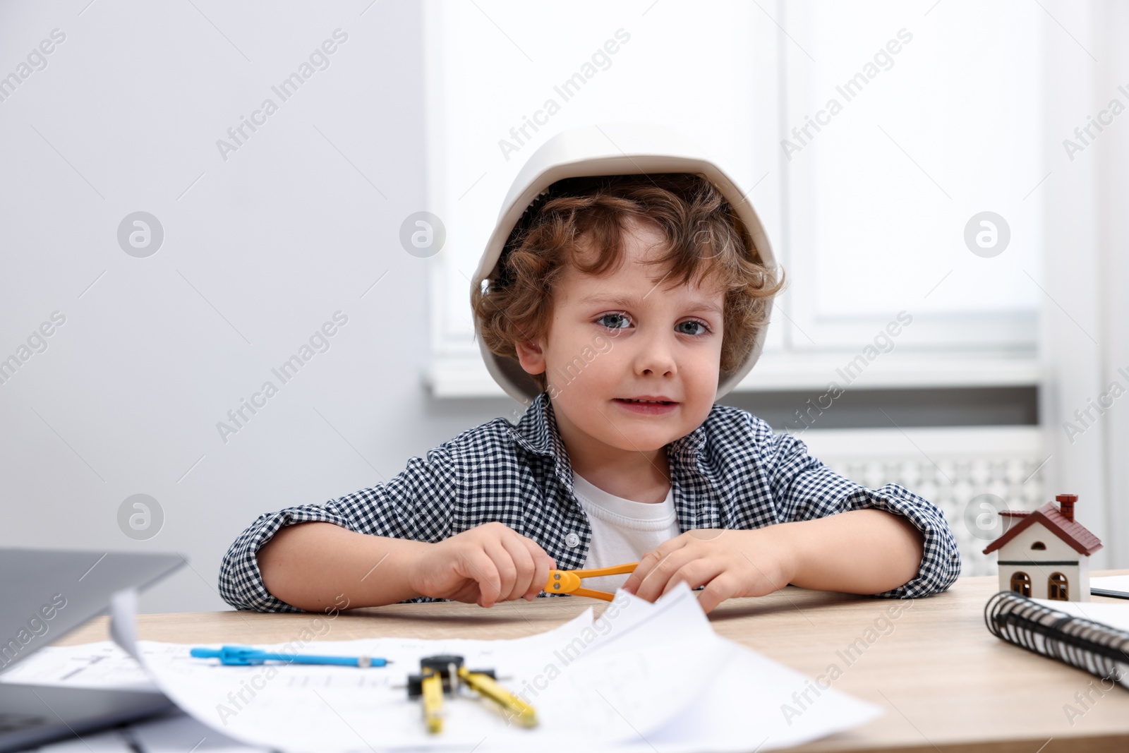 Choice of profession. Little kid in hard hat with divider pretending to be engineer at table indoors Photo of Choice of profession. Little kid in hard hat with divider pretending to be engineer at table indoors