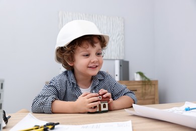 Photo of Choice of profession. Little kid in hard hat exploring house model at table indoors