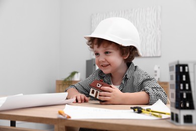 Photo of Choice of profession. Little kid in hard hat exploring house model at table indoors