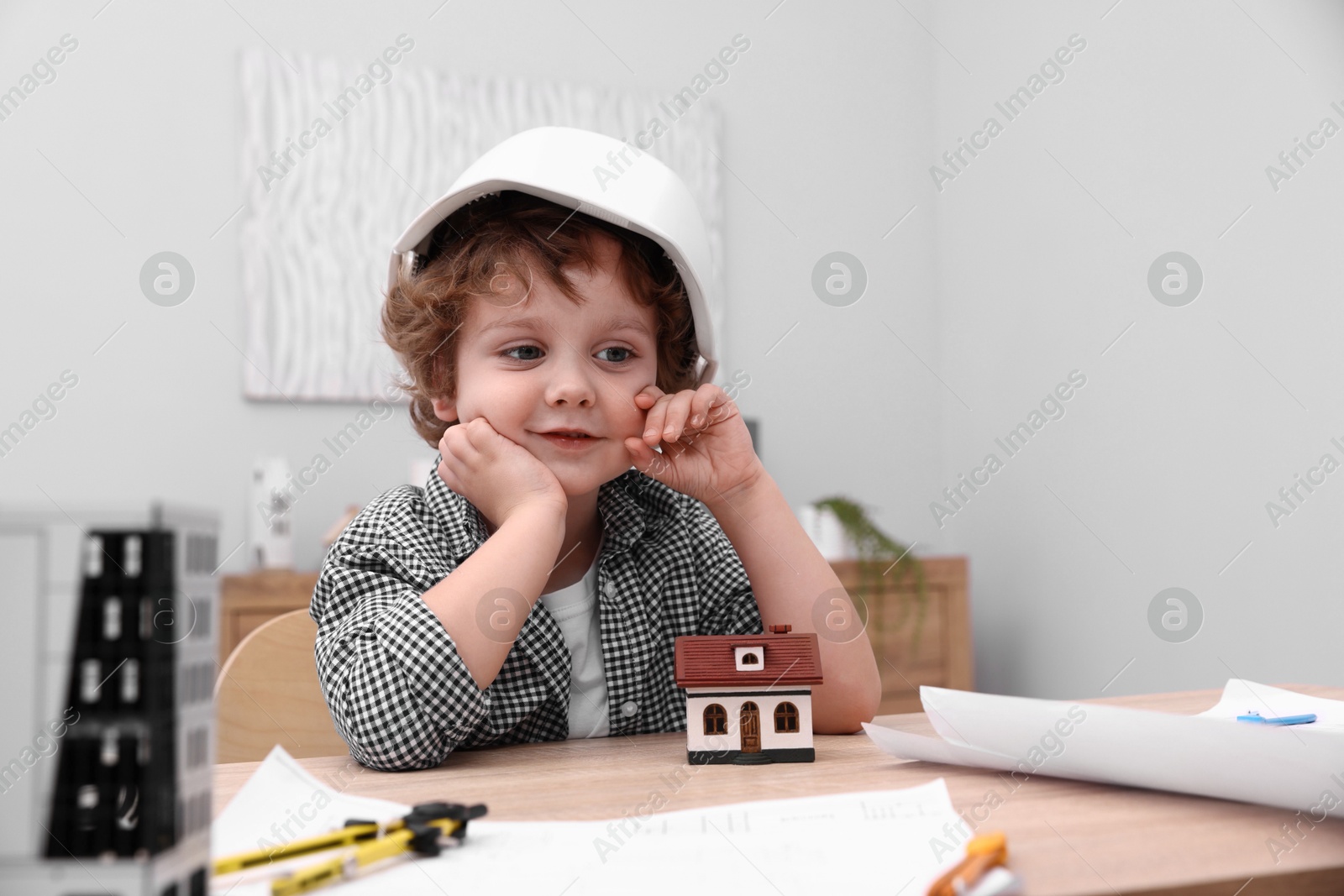Choice of profession. Little kid in hard hat exploring house model at table indoors Photo of Choice of profession. Little kid in hard hat exploring house model at table indoors
