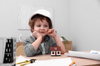 Photo of Choice of profession. Little kid in hard hat exploring house model at table indoors