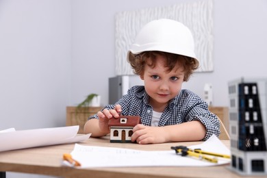 Photo of Choice of profession. Little kid in hard hat exploring house model at table indoors