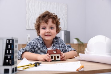 Photo of Choice of profession. Little kid in hard hat exploring house model at table indoors