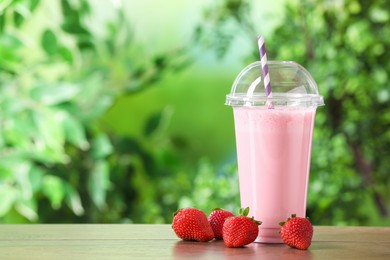 Photo of Tasty milkshake in plastic cup and strawberries on wooden table outdoors, space for text