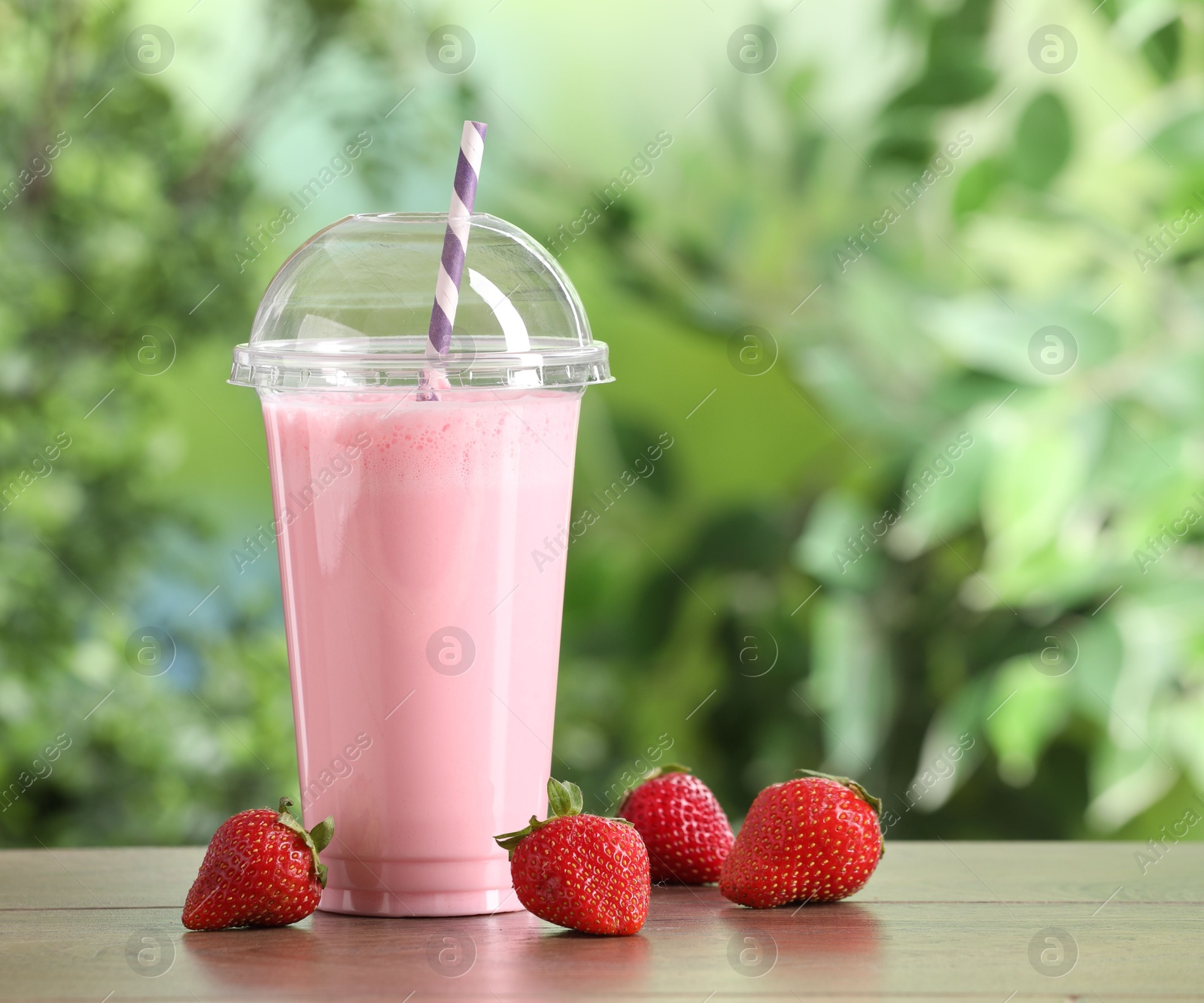 Photo of Tasty milkshake in plastic cup and strawberries on wooden table outdoors