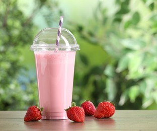 Photo of Tasty milkshake in plastic cup and strawberries on wooden table outdoors
