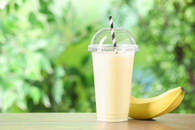Photo of Tasty milkshake in plastic cup and banana on wooden table outdoors, space for text