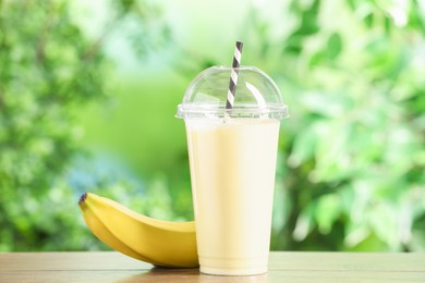 Photo of Tasty milkshake in plastic cup and banana on wooden table outdoors