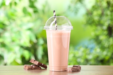 Photo of Tasty milkshake in plastic cup and chocolate on wooden table outdoors
