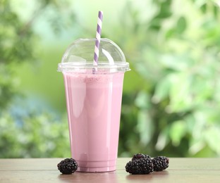 Photo of Tasty milkshake in plastic cup and blackberries on wooden table outdoors
