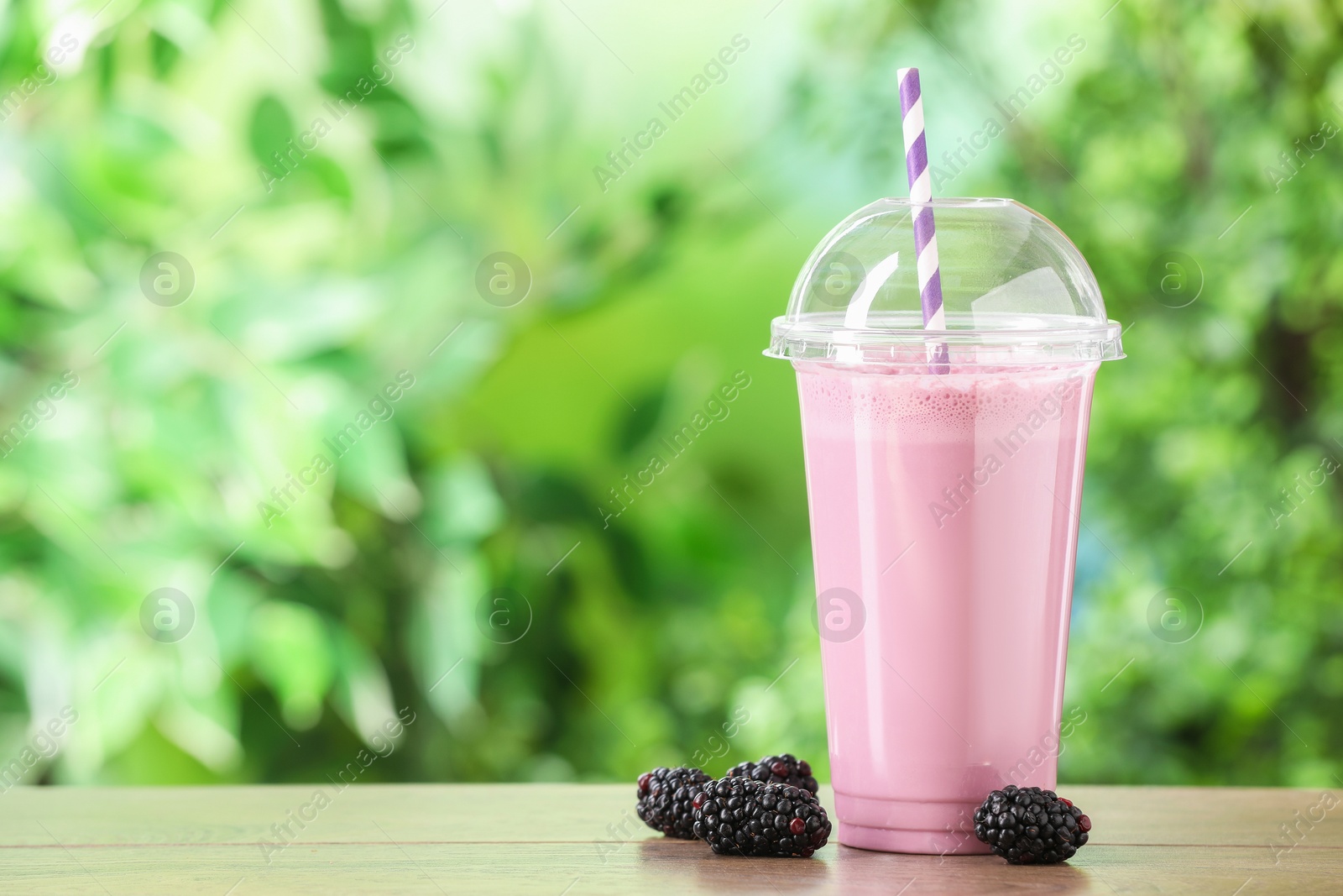 Photo of Tasty milkshake in plastic cup and blackberries on wooden table outdoors, space for text