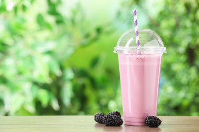 Photo of Tasty milkshake in plastic cup and blackberries on wooden table outdoors, space for text