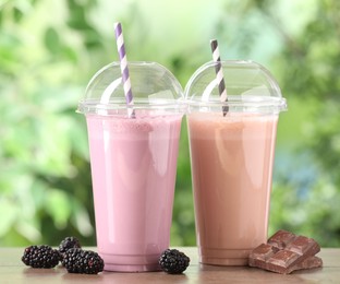 Photo of Tasty milkshakes in plastic cups and ingredients on wooden table outdoors